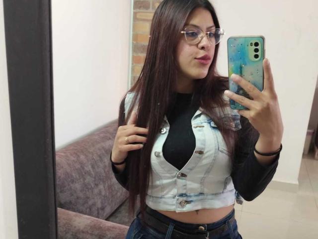 ElaiaaBakerr with long brown hair, glasses, and a denim vest over a black top, holding a phone with a blue case, showing her tits in a mirror selfie.