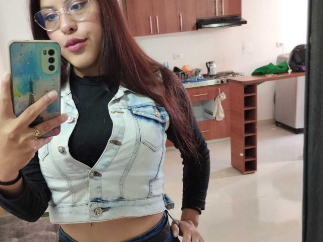 ElaiaaBakerr with long red hair, glasses, pink lipstick, black turtleneck, and light blue denim vest, showing her tits in a mirror selfie, holding a phone with a colorful case.