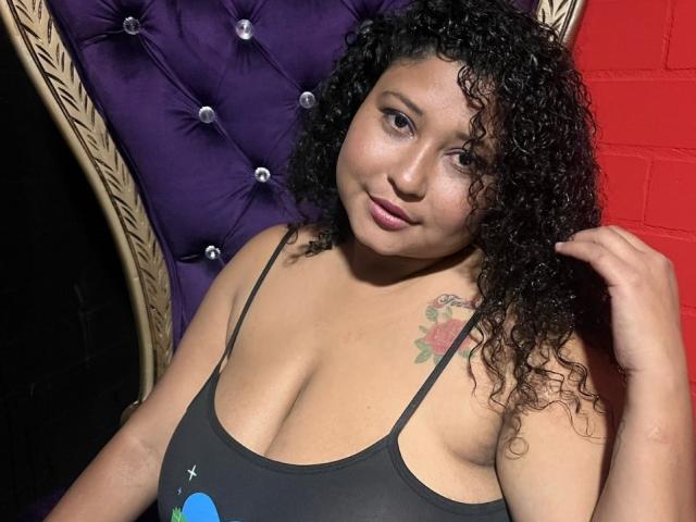 Curly-haired Ayliin with tan skin and a tattoo on her right shoulder, wearing a black tank top showing her big tits, sitting in front of a purple tufted chair.