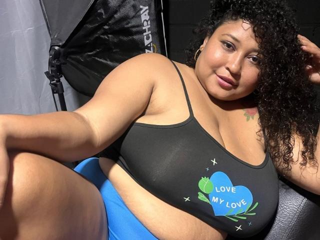 Ayliin with curly black hair, large breasts, and a black sports bra with a blue heart and "Love My Love" text, sitting and smiling at the camera.