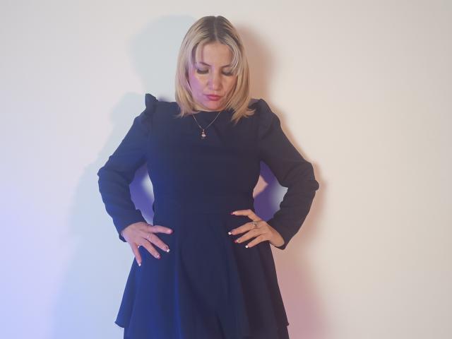 GiuliaAddams stands with hands on hips, wearing a black dress, blonde hair, and a necklace, looking down with a slight frown, her ass subtly highlighted by the dress.