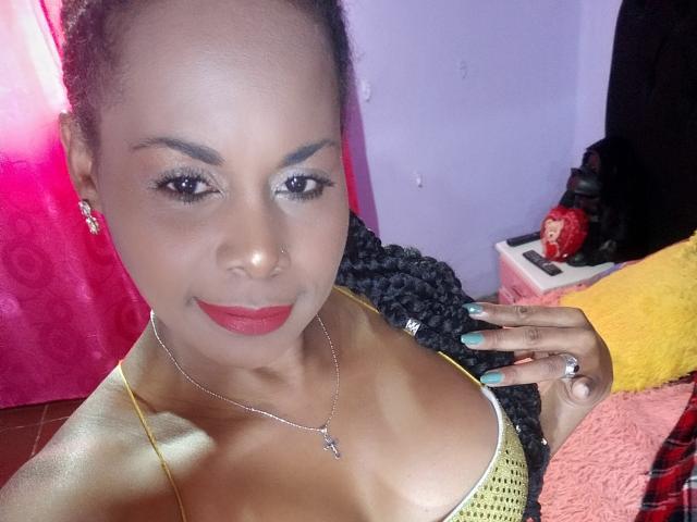 Afrodita Kane ha la pelle marrone scuro, rossetto rosso e unghie verdi, indossa una maglia gialla in rete che mette in mostra il seno, con una collana a forma di croce e un sorriso sicuro..