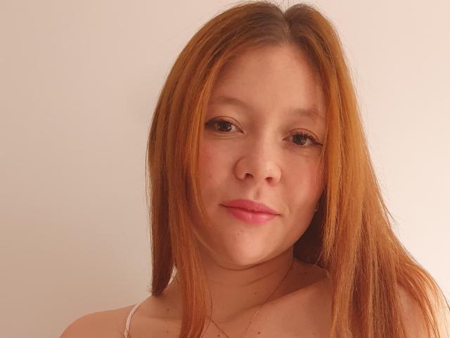 BlissfulEmberCouple has light brown hair, fair skin, and pink lips, looking directly at the camera with a subtle smile, wearing a white strap, and a simple gold necklace.