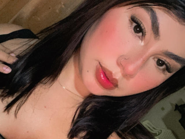 KimZhukova has shiny black hair, pink blush, red lipstick, and a nose ring. Her tits are barely visible, showing a black top. She has long eyelashes and a slight smile.