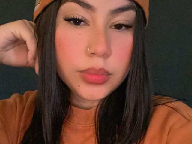 KimZhukova has dark hair, a nose ring, and is wearing a brown beanie. She's pouting with pink lips, half-closed eyes, and wearing a brown shirt.
