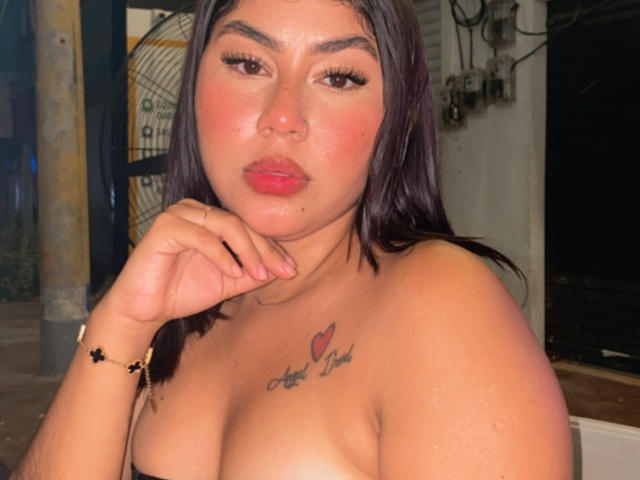 KimZhukova with dark brown hair, full pink lips, and a heart tattoo on her chest, her hand near her face, showing her bare tits, wearing a black bracelet.