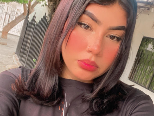 KimZhukova has dark hair, bold eyebrows, pink blush, and pink lipstick, with a black shirt, and a nose ring, looking at the camera with a slightly pouty expression.