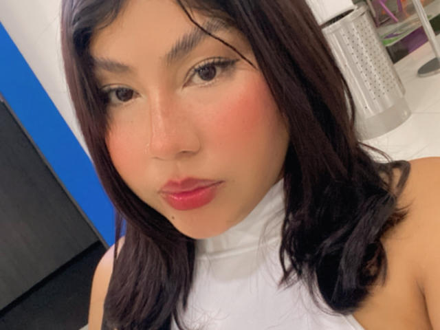 KimZhukova with dark brown hair, pink blush, red lipstick, and white top, showing a cute, slightly flushed face and looking at the camera with a subtle smile.