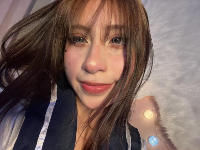 LilithAsian has brown hair, green eyes, and a slight smile with pink lips. She's wearing a dark blue top with white straps. Small moles on her cheeks. Soft lighting.