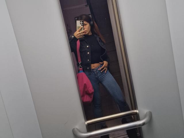 RinaQueenie with dark brown hair, black crop top, blue jeans, pink bag, and black jacket, showing her small ass in a mirror selfie, hand on hip.