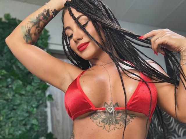 NadiaSpark with long braids, red bikini top, red lipstick, tattoo under boobs, holding hair, arm tattoo, necklace, confident smile, tan skin, sexy, room with green leaf wall.