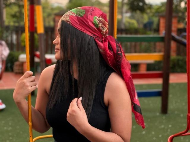 MiiaPalmer with long black hair, wearing a red bandana, black tank top, holding a yellow swing bar, looking to the side, in a playground, with a calm expression.