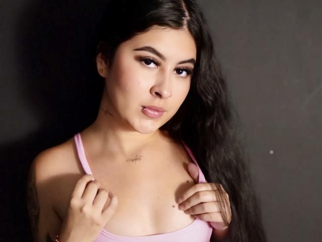 NicoleCollins with long dark hair, pink tank top, holding her small tits, subtle tattoo on chest, slightly parted pink lips, dark background, soft light, focused expression.