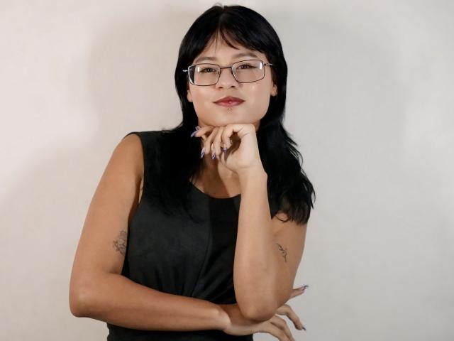 ElizabethRoux with black hair, glasses, and a sleeveless black top, resting her chin on her hand, showing tattoos on her arms, and smiling subtly.
