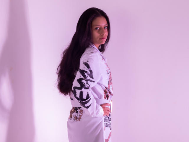 AteneaSexy69 with long dark hair, wearing a white shirt with black graffiti-style text, standing side-profile, hands on hips, looking over shoulder, in soft purple light.