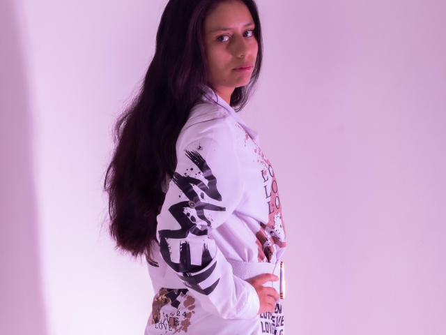 AteneaSexy69 with long black hair, white hoodie, and graphic prints, standing sideways, looking over shoulder, hands on hip, purple light, white background. Boobs not visible, pussy not visible.