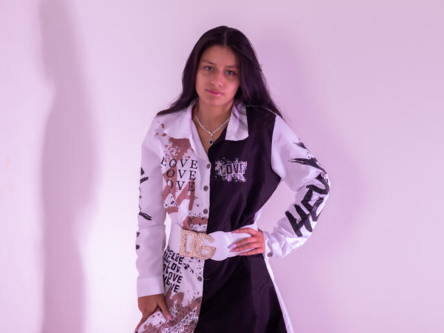 AteneaSexy69 with long black hair, wearing a white and black shirt with bold text and patterns, hands on hip, necklace, slight purple light, shadow on white wall.