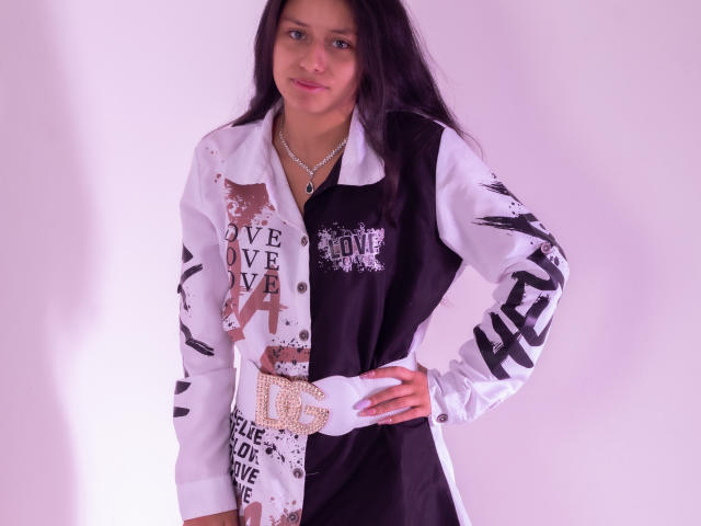 AteneaSexy69 with long black hair, pinkish skin, and small tits, wearing a black and white jacket with bold black and white graffiti, standing with hands on hips.