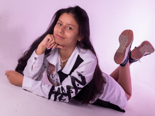 AteneaSexy69 with long dark hair, lying on her stomach, wearing a white shirt with black graffiti, black shoes, and a necklace, smiling, with her hand under her chin.