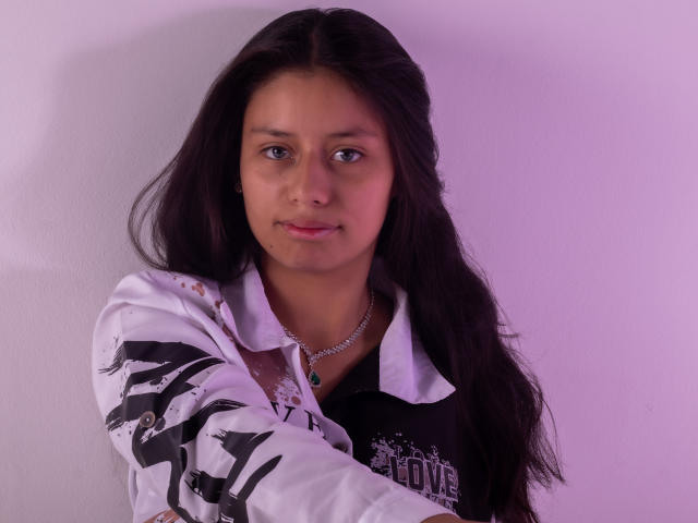 AteneaSexy69 has long black hair, brown eyes, and a cute face. She wears a white and black shirt with bird patterns, and a silver necklace with a blue pendant.