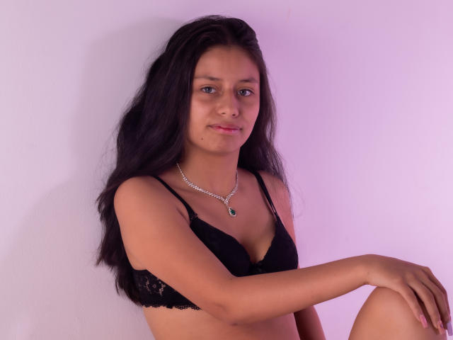 AteneaSexy69 with long black hair, wearing a black lace bra, silver necklace, and a slight smile, has small tits and a cute face, sitting with one arm resting.