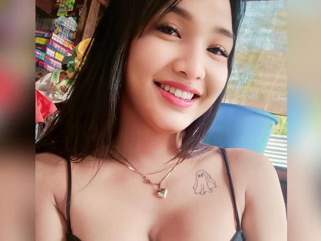 PinaySophia with black hair, smiling, wearing a black spaghetti strap top, showing small tits, a heart necklace, and a ghost tattoo on her right chest.