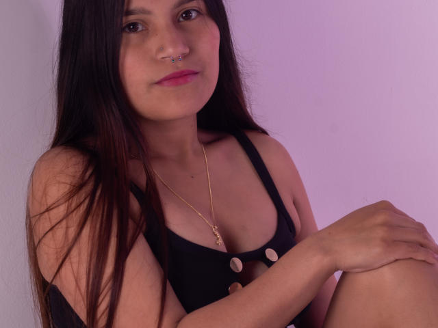 MelanieGates with long brown hair, nose ring, black tank top with buttons, small tits, gold necklace, sitting with knees up, pinkish light, slight smile, relaxed, cute.