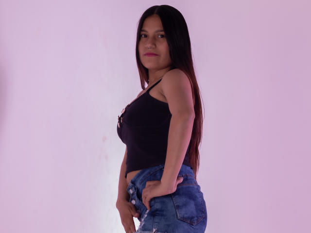 MelanieGates with long black hair, wearing a black tank top showing her tits, and blue jeans, standing sideways, hands in pockets, pinkish background, serious expression.