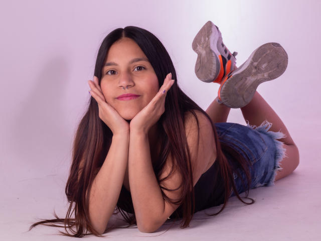 MelanieGates with long brown hair, black tank top, denim shorts, and orange sneakers, lying on her stomach, hands under her chin, smiling, ass up, feet in the air.