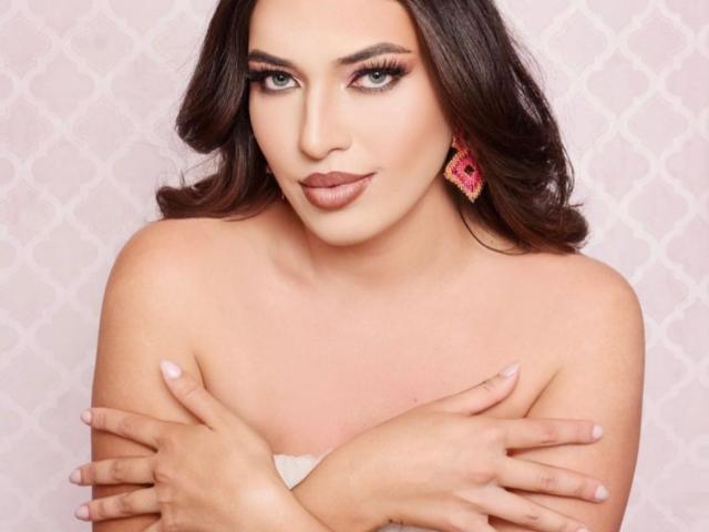 RachelWildest has brown hair, blue eyes, and is crossing her hands over her tits, wearing pink earrings with a slight smile. She has a fair skin tone and pink lipstick.