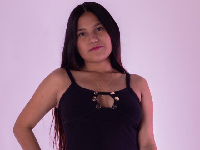 MelanieGates has long dark hair, light skin, and wears a black tank top with metal ring cutouts, a gold necklace, and a pink nose ring, standing with hands on hips.