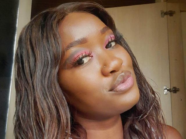 TastyDreamGirl has dark brown skin, long wavy hair, pink eyeshadow, thick eyelashes, a nose piercing, and glossy pink lips, smiling slightly with a subtle pout.