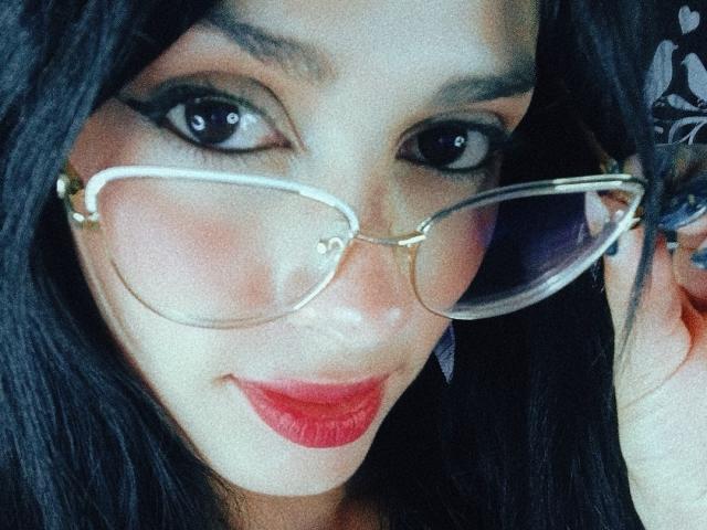 AnaisHilton with glasses, dark eyes, and red lipstick, smiling at the camera, holding her glasses with black hair covering her ears. Her tits are not visible.