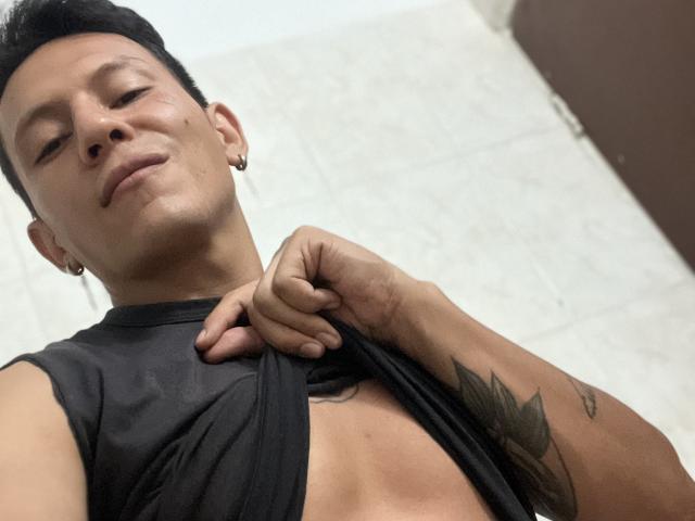 Michael Strong, com a pele morena, cabelo preto curto, pequenas argolas nos ouvidos, tatuagem no antebraço direito, levantando a camisa preta para mostrar o peito plano e os músculos abdominais, sorrindo levemente, olhando para baixo..