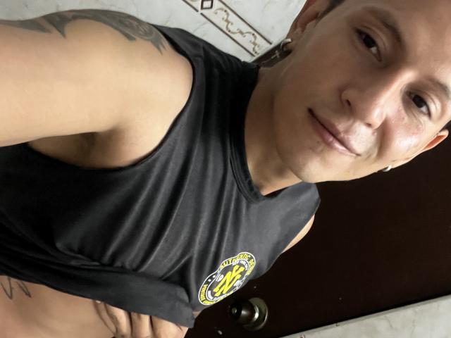 MichaelStrong with a shaved head, wearing a black sleeveless shirt, showing a tattoo on his right arm, and a subtle smile, holding his shirt up to reveal his belly.