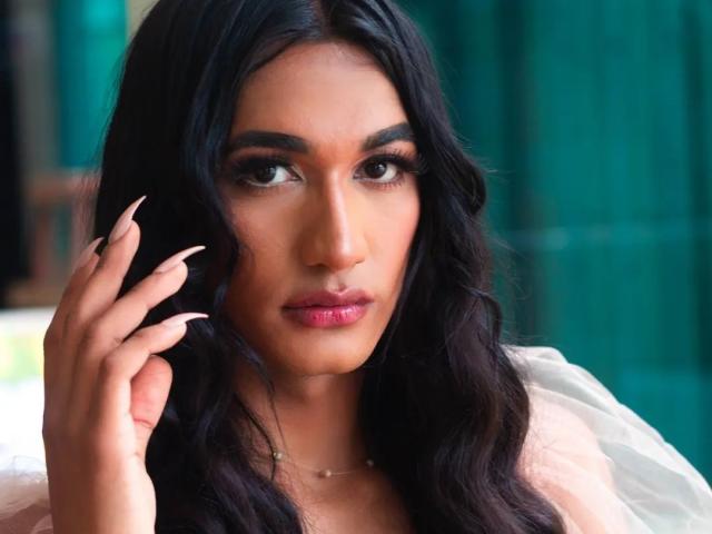 Alexa69s has long black wavy hair, dark eyes, and pink lipstick. She's touching her hair with long, pointed nails, wearing a white top, looking serious.