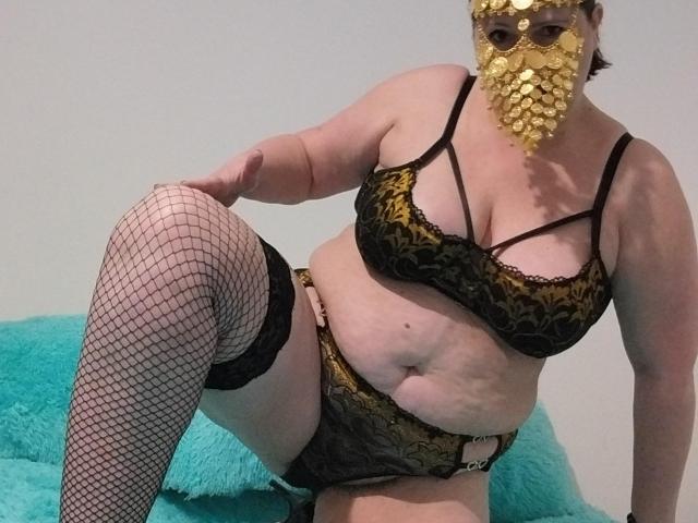 LiliChattePoilue, de talla grande, con una máscara de pan de oro, un sujetador de encaje negro y tanga, medias de red hasta el muslo, sentada sobre un cojín turquesa, mostrando sus pechos y su vientre, curvilínea y sexy..