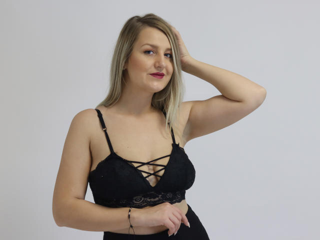 Maya Trainor hat blondes Haar, helle Haut und kleine Brüste. Sie trägt einen schwarzen Spitzen-BH mit Kreuzungsriemen und steht mit einem Arm erhoben da, leicht lächelnd..