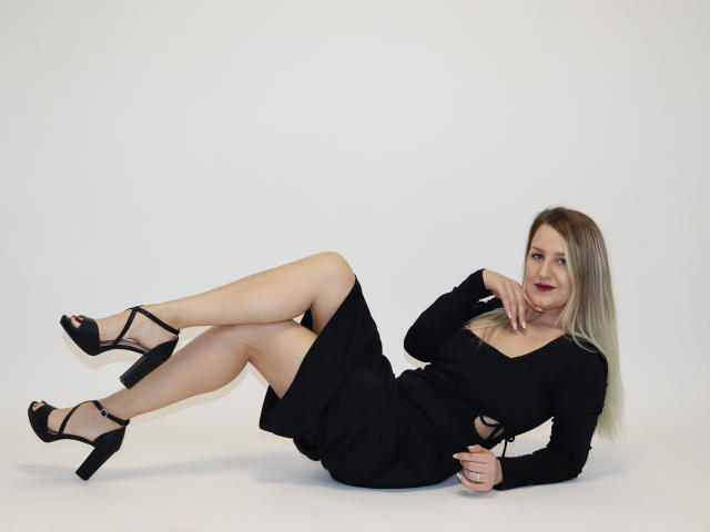 Maya Trainor mit blonden Haaren, einem schwarzen Kleid und High Heels, liegt auf dem weißen Boden, die Beine angezogen, die Finger in der Nähe des Mundes, und zeigt ihren nackten Hintern und ihre Brüste. Sie lächelt..