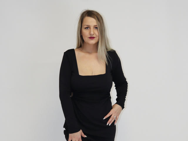 MayaTrainor has blonde hair, black dress with a square neckline, showing her tits, standing with one hand on her hip, white background, red lipstick, and a confident smile.