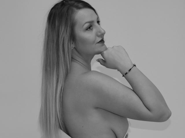 Schwarz-weißes Foto von Maya Trainor mit langen blonden Haaren, im Profil, Hand in der Nähe des Mundes, kleine Ohrringe, Armband, freie Schultern, die über die Schulter schaut und dabei ihre Brüste zeigt..