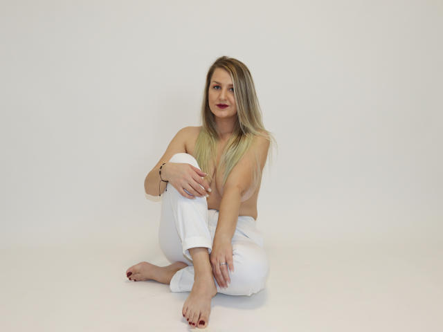 MayaTrainor, topless, with long blonde hair, sitting on white floor, legs crossed, white pants, bare feet, red toenails, small tits, light skin, subtle smile, minimal makeup, ring on right hand.