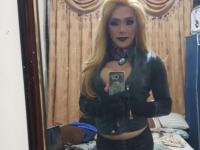 QueenOfPainSasha with blonde hair, black leather top, black gloves, purple necklace, dark lipstick, holding phone, showing large tits, standing in front of mirror, reflecting herself.