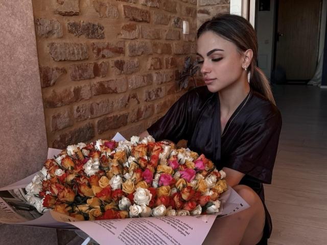 KarolinaSykes with her tits down, wearing a black satin robe, holding a bouquet of colorful roses. She's got brown hair in a ponytail, and her eyes are closed.