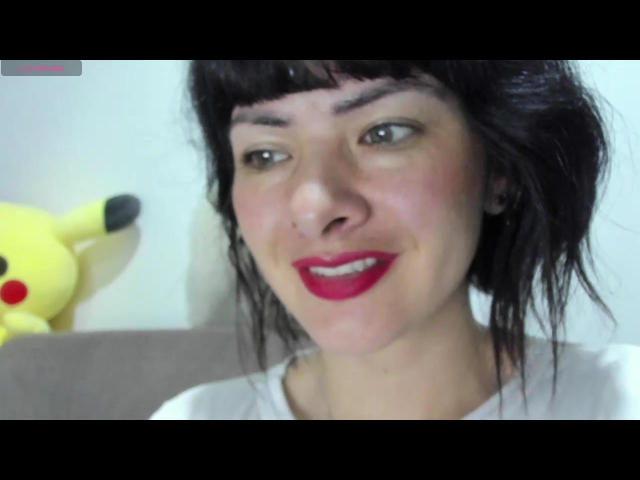 Antomouth has dark, messy hair, red lipstick, and is smiling with a cute Pikachu toy on the left. She's wearing a white shirt and has small earrings.