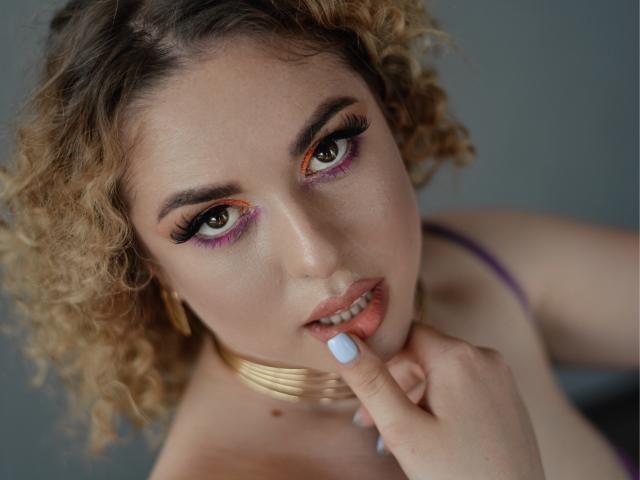 ArishkaLulu has curly blonde hair, purple eyelids, and white lipstick, with a finger on her pink lips, wearing a gold necklace and purple top, looking seductively at the camera.