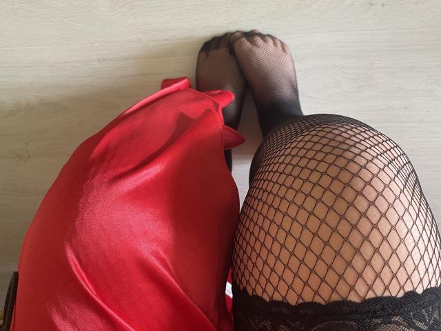 Las piernas de Madrinia, vestida con un vestido de satén rojo y medias de rejilla negras, con los pies tocándose, sobre un suelo de madera blanca. Los senos no son visibles, el enfoque está en las piernas y las medias..