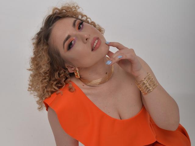 ArishkaLulu, com cabelos loiros cacheados, vestido laranja, colar e pulseira dourados, tocando o queixo, sombra rosa nos olhos, unhas azuis e um sorriso sedutor, mostrando os seios..
