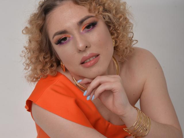 ArishkaLulu with curly blonde hair, orange eyeshadow, pink lipstick, gold hoop earrings, and orange top, showing off her tits and light blue nails, looking sexy and confident.
