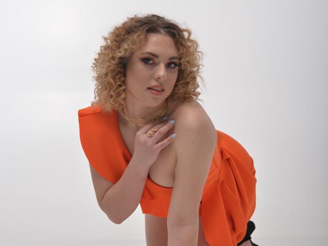 ArishkaLulu with curly blonde hair, wearing an orange dress, bending forward, showing her tits, light makeup, gold necklace, and blue nails, looking at the camera.